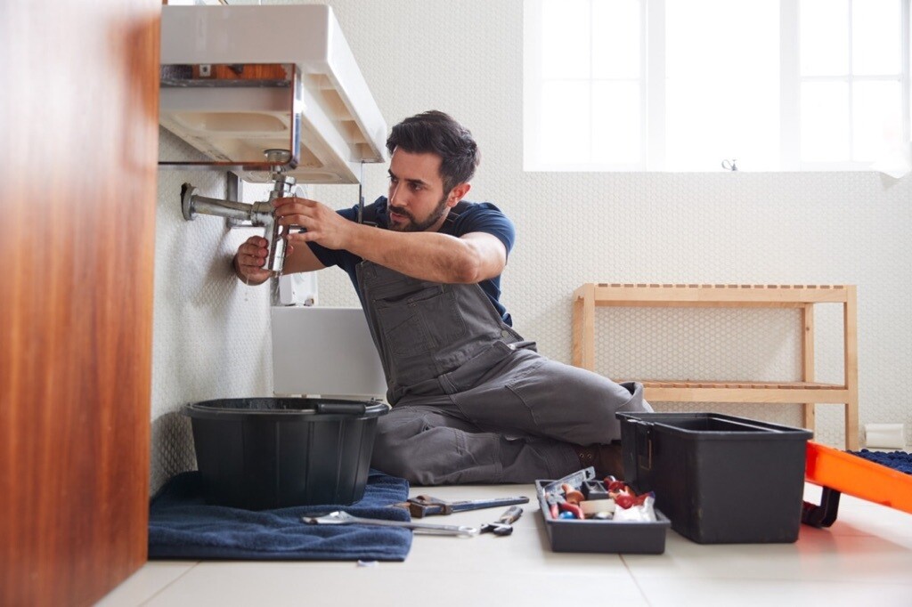 Person wearing overalls while sitting on the ground fixing pipes under a sink